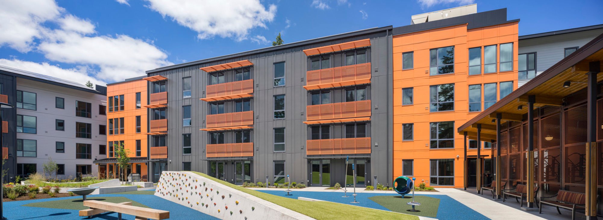 A panoramic view of the Wynne Watts apartment complex on a sunny day from the perspective of the interior courtyard, with a children's play area in the foreground. 