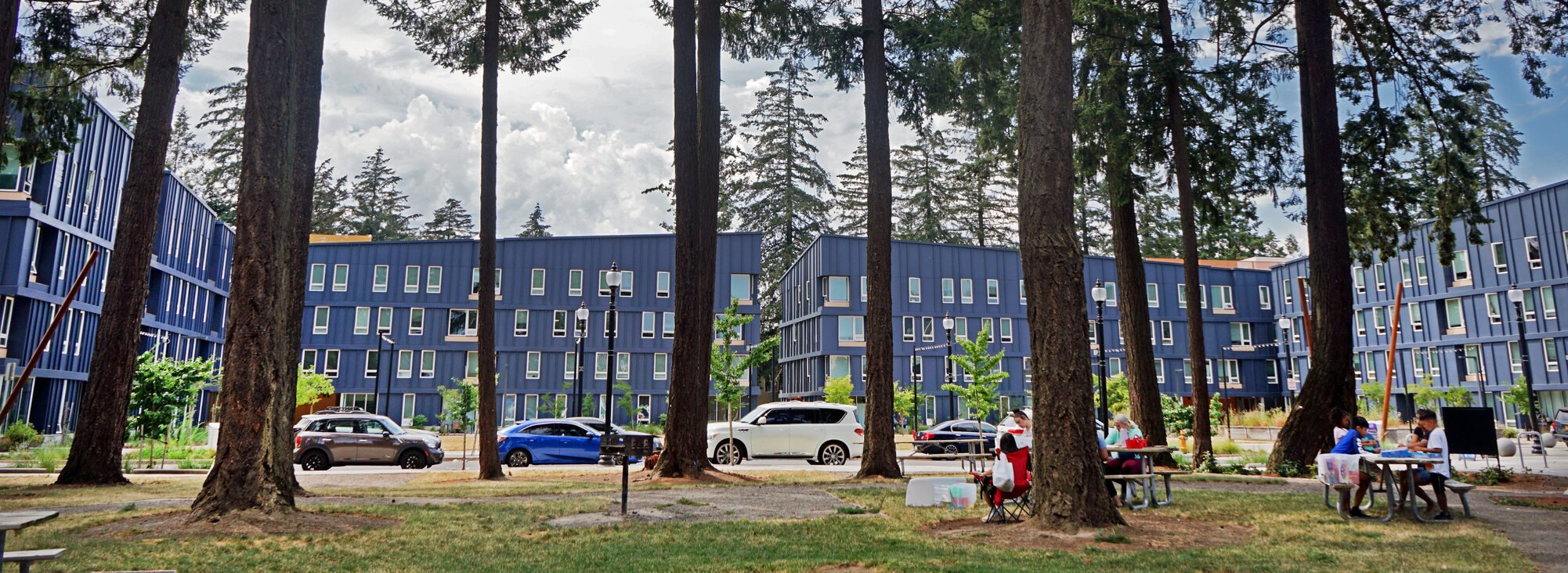 Housing complex with dark blue exterior and tall pine trees in the foreground. Two picnic tables with people are on the lower right.
