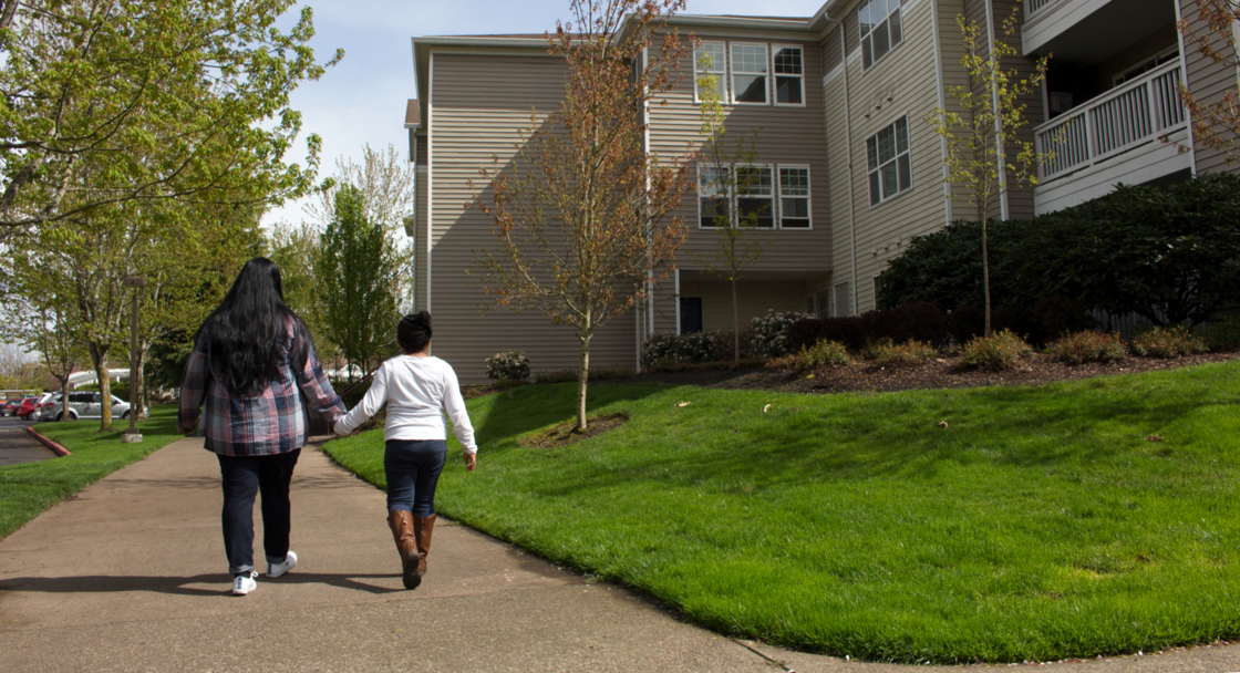 an adult and a child walk hand-in-hand on a sidewalk in front of an apartment complex