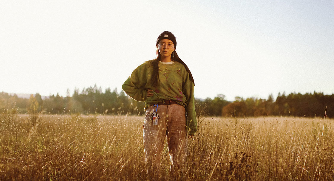 A 20-something young person with long black hair stands in a field of dried grass that comes up to their waist.