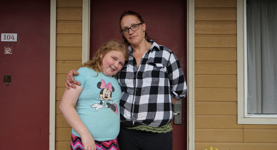 A woman wearing glasses and a black-and-white plaid shirt stands with a child wearing a Minnie Mouse t-shirt and pink striped pants in front of the entrance door to an affordable housing unit