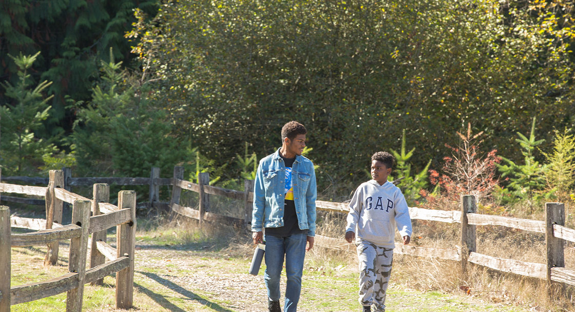 A young Black man and a Black child talk as they walk along a fenced trail in a park.