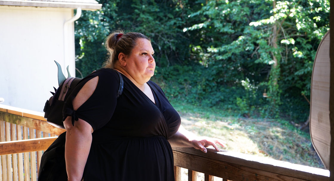Woman standing at a wooden railing looking into the distance, wearing a black dress.