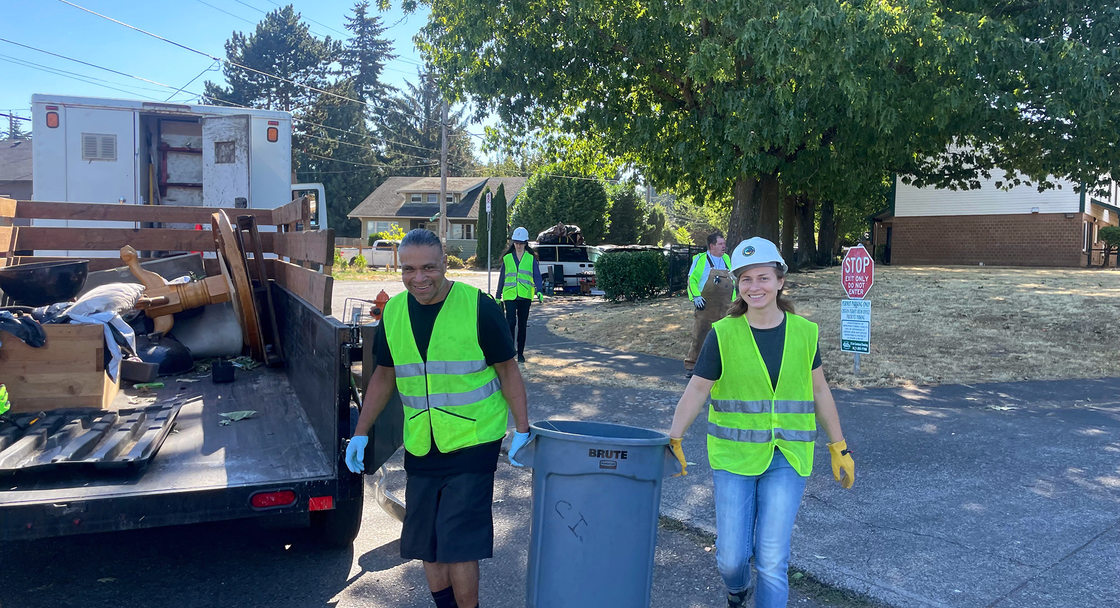A man and woman carry a trash can to an open trailer bed. Both wear neon yellow high visibility vests.