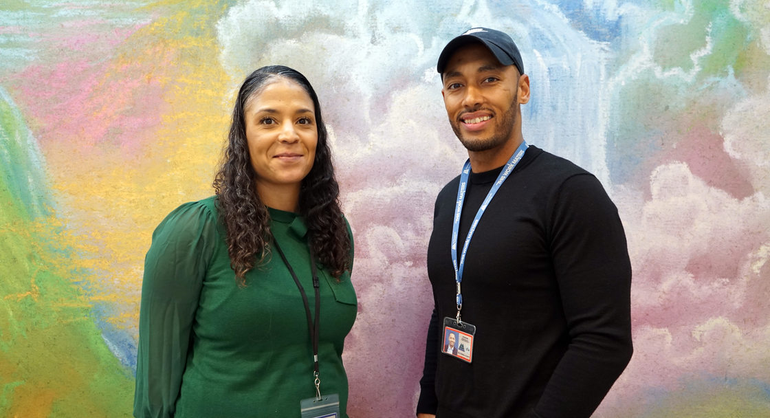 A Black man and woman standing in front of a colorful pastel mural, smiling