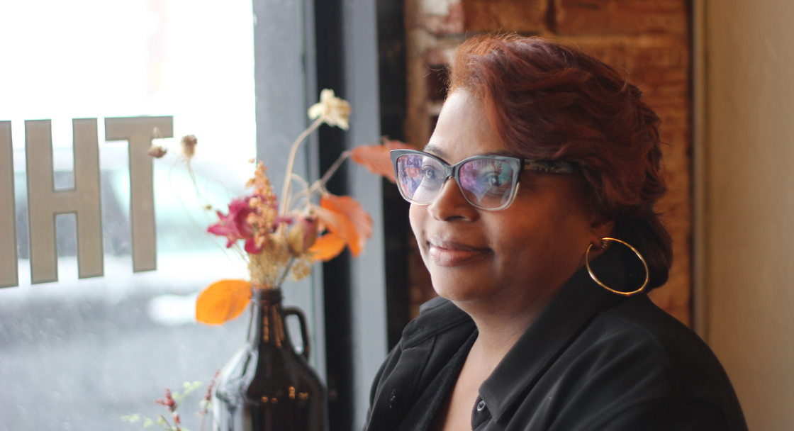 Close up of a woman with cat eye glasses, hoop earrings and a black blazer with auburn hair smiling. A window and vase with flowers are in the background
