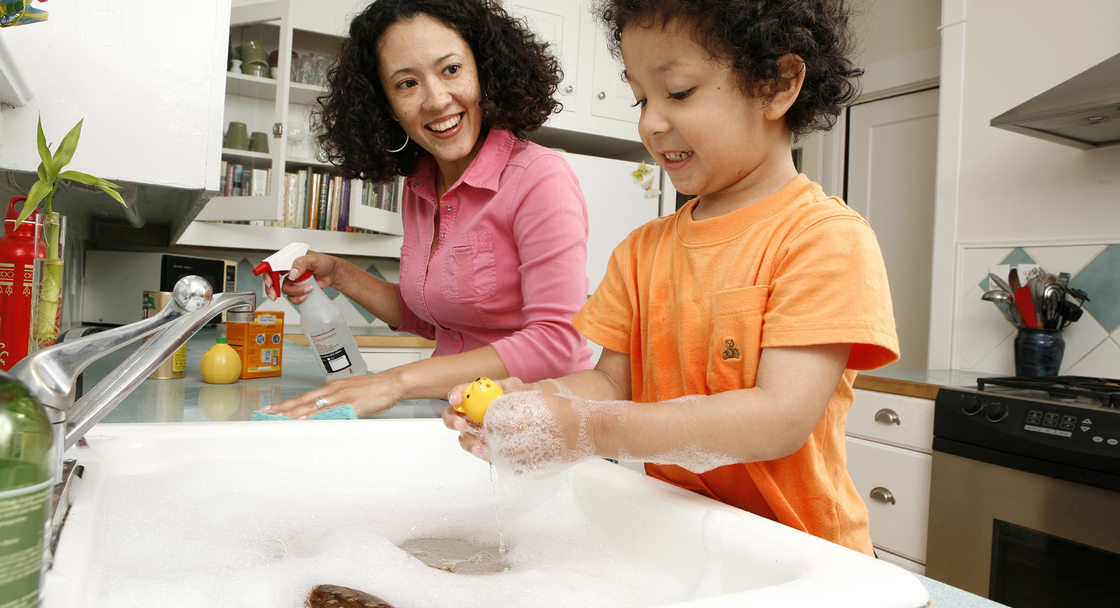 A woman and her son lean over a kitchen counter. The son plays with bubbles in the sink while the mom sprays down the countertops