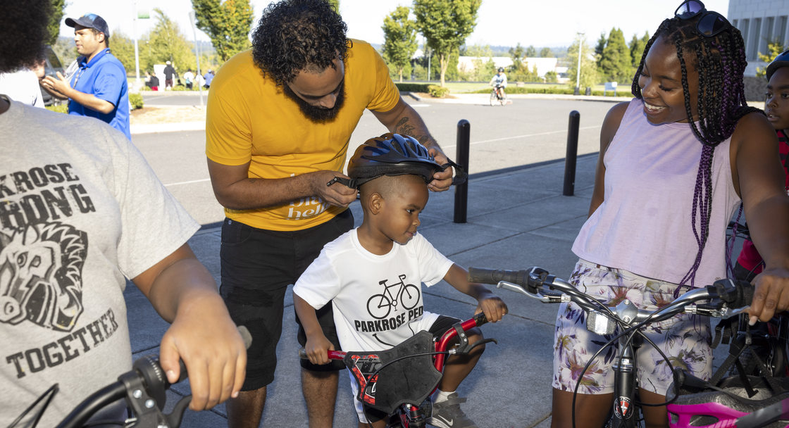 Image of an adult in a yellow t-shirt putting a bike helmet on a child who is sitting on a bike.