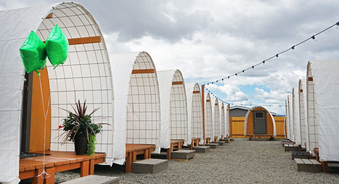 Row of structures with curved white roofs and wooden porches on a gravel lot. 