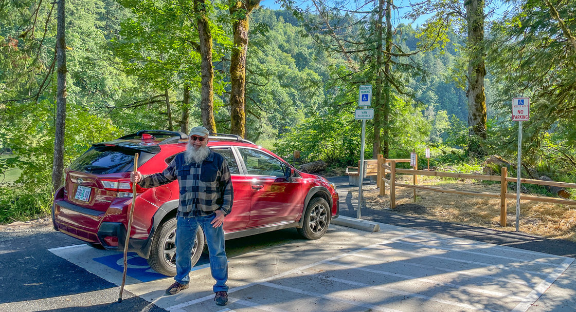 Len Otto stands in front of his red SUV that is parked in a marked accessible parking spot. Otto is using a cane. The spot is surrounded by trees.  