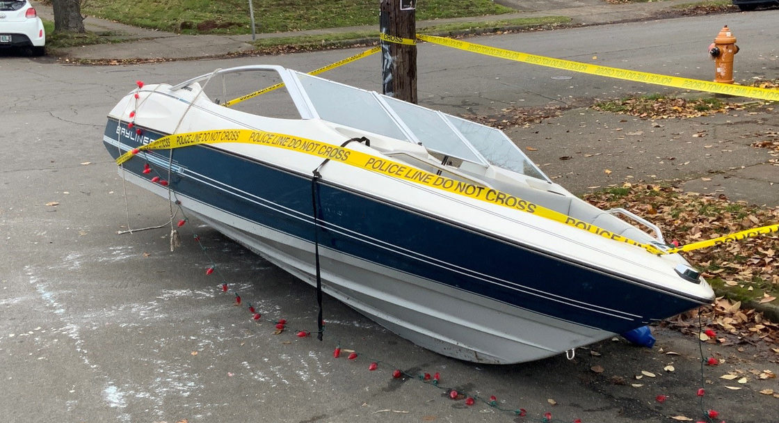 A white boat with a navy blue strip leans over on a city street. Yellow police tape marks the edge of the boat and a light pole behind it.