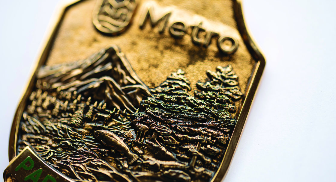 A close up of a bronze park ranger badge has details of mountains, trees and a river. It reads "Metro Park Ranger". 