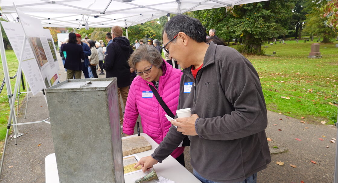 woman and man standing at a table under a canopy in a cemetery, looking at Chinese writing on a gravestone lying on the table