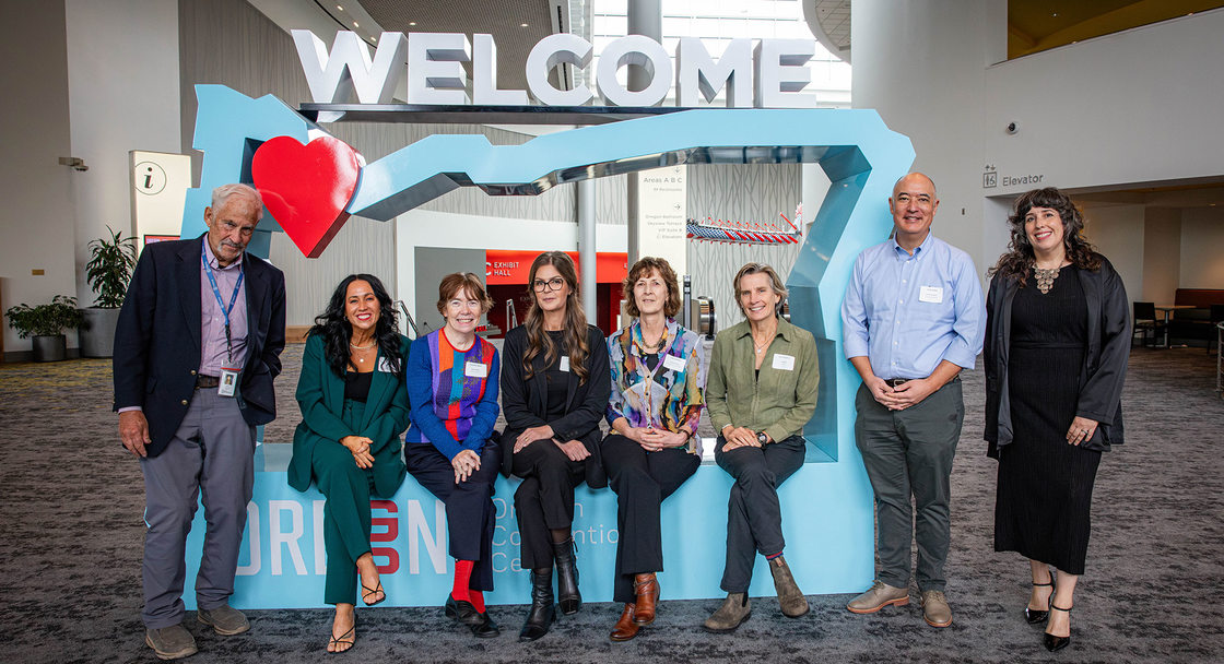 8 people are infront of a light blue cutout of Oregon, 5 are sitting in the cutout, 3 are standing