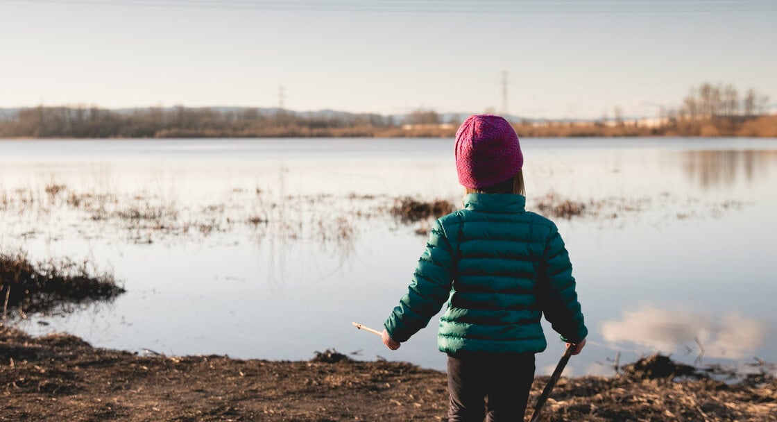 A small child faces away from the camera at the shore of a lake on a sunny winter day.