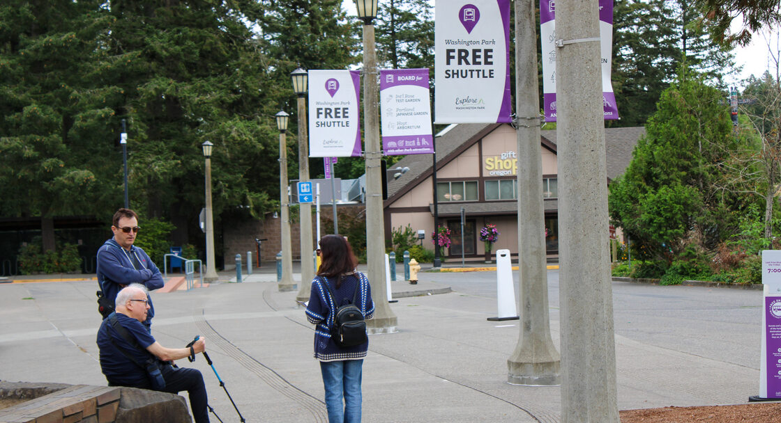 Three people, standing and sitting, wait on the sidewalk for the Washington Park free shuttle. Signs for the free shuttle hang from light posts that line the sidewalk and trees surround the area.