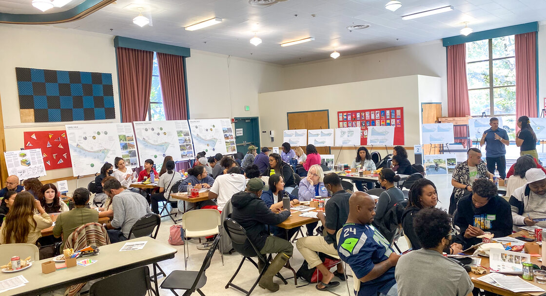 A community center room is filled with about 50 people sitting around folding tables.
