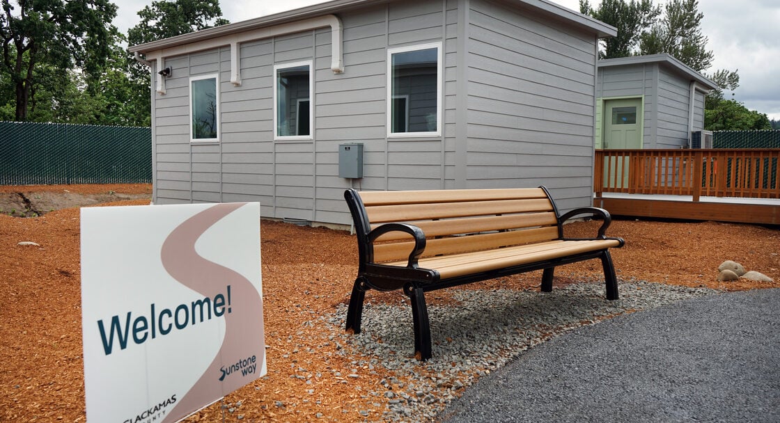 Bench in front of a grey bungalow, with a Welcome sign next to it. 