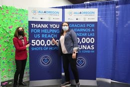 Governor Kate Brown and another woman wear masks and stand next to a banner for All4Oregon at the Oregon Convention Center mass COVID-19 vaccination site