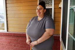 Woman in grey shirt with a bun smiling and holding a cane, standing in front of a marigold colored wall