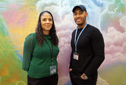 A Black man and woman standing in front of a colorful pastel mural, smiling