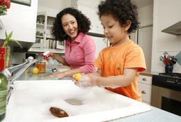 A woman and her son lean over a kitchen counter. The son plays with bubbles in the sink while the mom sprays down the countertops